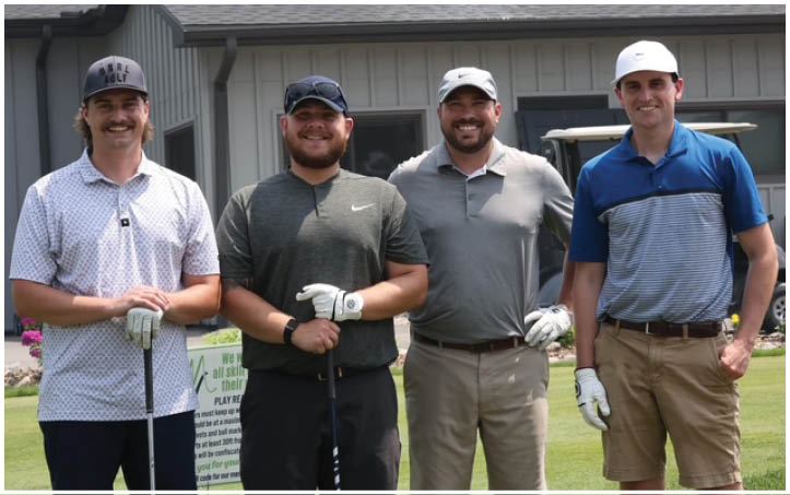 Four adults wearing golf attire and gloves stand side by side smiling on a golf course with a building and golf cart in the background. Description generated by AI