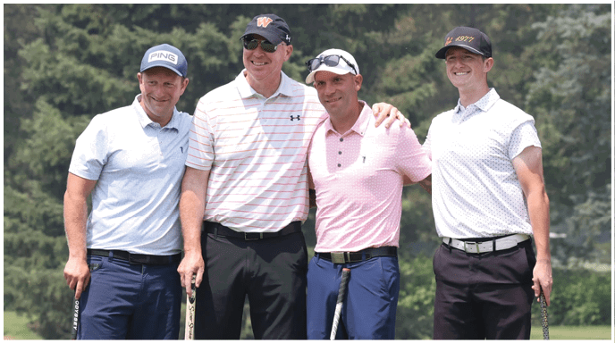 Four adults wearing golf attire stand closely together on a golf course holding golf clubs and smiling. Description generated by AI