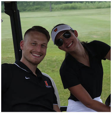 Two adults smiling and sitting together in a golf cart on a grassy area, with one wearing a visor and sunglasses and the other wearing a black polo shirt. Description generated by AI
