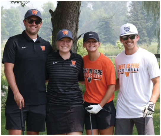Four people wearing sports apparel and hats are smiling outdoors near a tree and grassy area, with two holding golf clubs. Description generated by AI