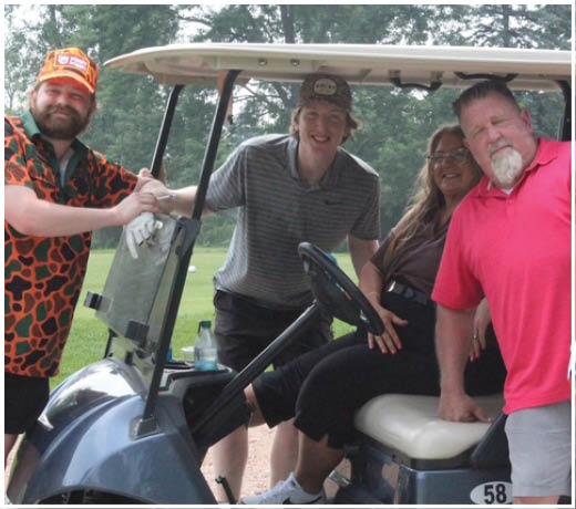 Four people smiling and posing together around a golf cart on a grassy outdoor area. Description generated by AI