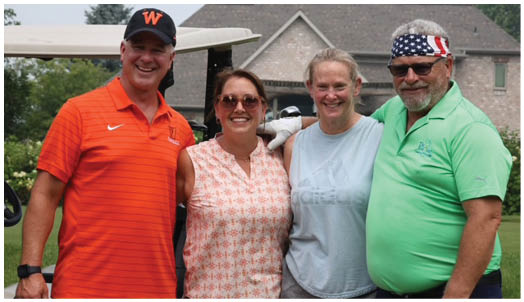 Four adults wearing casual clothing are standing closely together outdoors with a golf course and building in the background. Description generated by AI