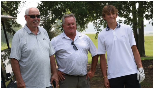Three people stand outdoors in casual golf attire with trees and a grassy area in the background. Description generated by AI