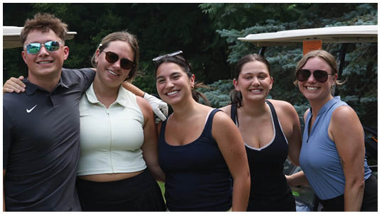 Five people wearing casual sporty clothing and sunglasses are standing closely together outdoors with arms around each other, smiling in front of a golf cart. Description generated by AI