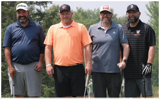 Four adults wearing casual golf attire and hats stand side by side on a golf course holding golf clubs with trees in the background. Description generated by AI