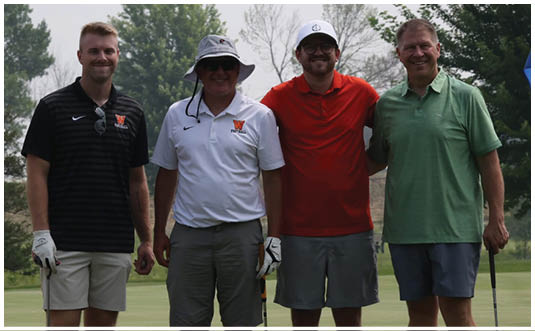 Four adults wearing casual golf attire stand together on a golf course near a hole with a blue flag. Description generated by AI