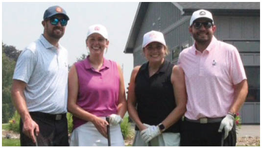 Four adults wearing golf attire stand on a golf course holding golf clubs with a building and greenery in the background. Description generated by AI