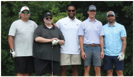 Five adults wearing casual golf attire stand together on a golf course with trees in the background, some holding golf clubs. Description generated by AI