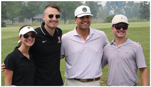 Four adults wearing casual golf attire and sunglasses stand closely together smiling on a golf course. Description generated by AI