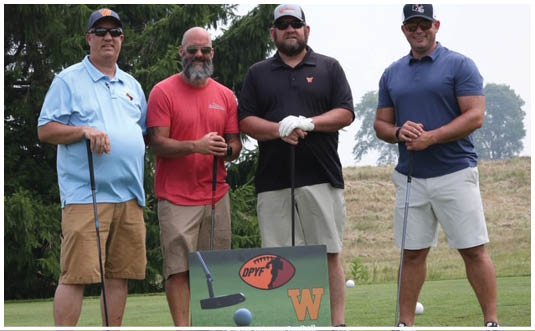 Four adults wearing casual golf attire stand on a golf course holding golf clubs next to a sign with a golf-themed logo and the letter W. Description generated by AI