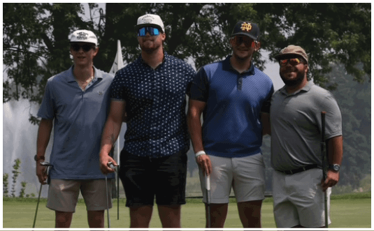 Four adults wearing casual golf attire and hats stand on a golf course holding golf clubs with trees and a body of water in the background. Description generated by AI