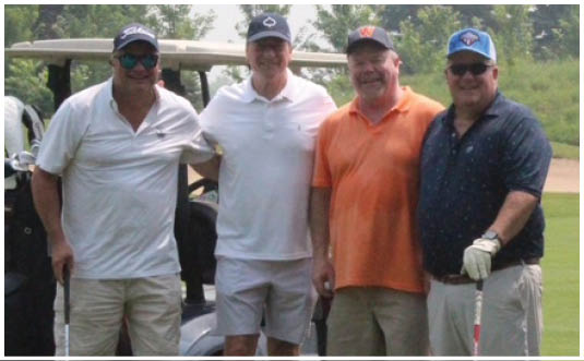Four adults wearing casual golf attire stand on a golf course with golf carts and clubs in the background. Description generated by AI