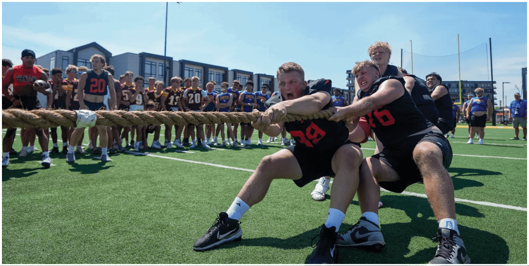 A group of people wearing sports jerseys are engaged in a tug-of-war on a grassy field with others watching in the background. Description generated by AI