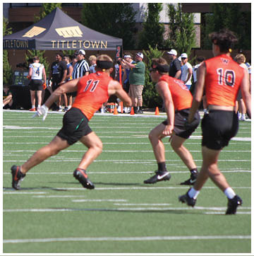 People wearing sports uniforms are playing a game on a green field with a tent labeled “Titletown" and spectators in the background. Description generated by AI