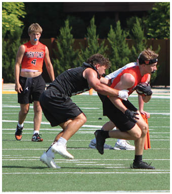 Two adults wearing athletic clothing are engaged in a competitive running or football drill on a grassy field with a referee and another person in the background. Description generated by AI