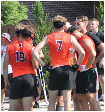 A group of people wearing orange athletic jerseys and black shorts are standing together on a sports field, some with headbands and one using crutches. Description generated by AI