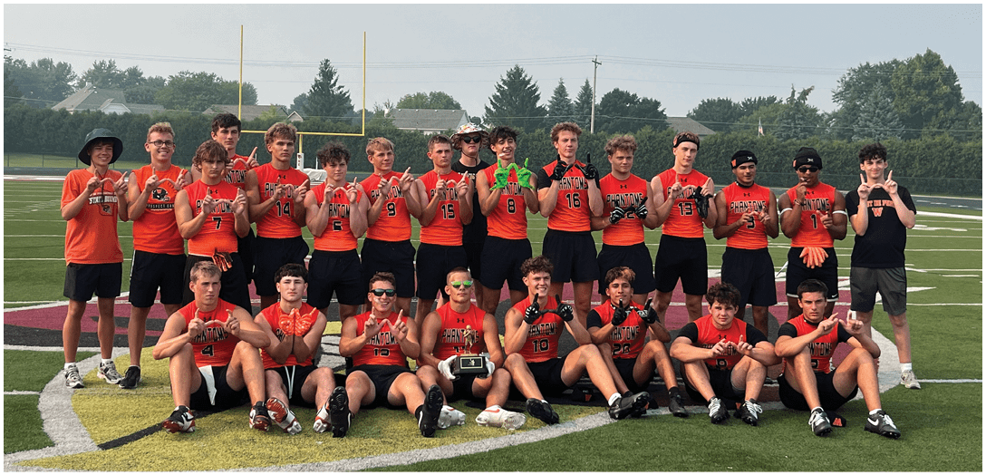 A group of people wearing orange sports jerseys stand on a football field holding up their fingers in a celebratory gesture. Description generated by AI