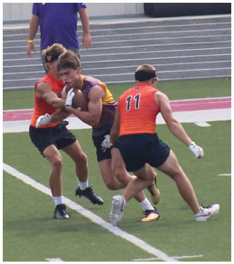Three people are playing football on a field with one person in a maroon and yellow jersey holding the ball while two others in orange jerseys attempt to block or tackle, and a person in a purple Vikings shirt and yellow cap watches from the sidelines. Description generated by AI