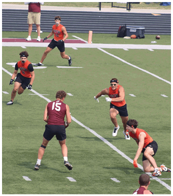 Five adults wearing athletic clothing are actively playing a sport on a green field with white lines, with one person in a maroon shirt facing four others in orange shirts. Description generated by AI