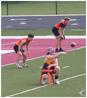 Three adults wearing athletic clothing are positioned on a sports field, with two standing on a red track area and one running on the green turf. Description generated by AI