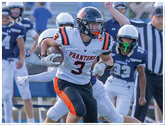 A person wearing a black and orange football uniform with the word Phantoms is running with the ball while being tackled by a person in a white uniform on a football field with other players and a referee in the background. Description generated by AI