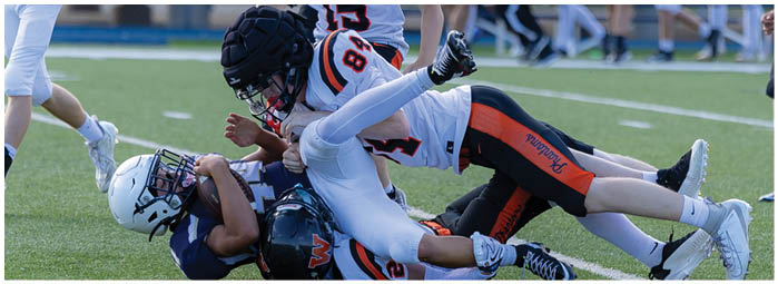A person in a football uniform with the number 15 is helping another person in a football uniform lying on the ground on a sports field with other players and officials in the background. Description generated by AI
