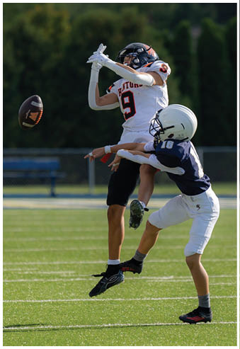Two people in football uniforms are engaged in a play on a grassy field, with one person jumping and the other reaching up to block or catch. Description generated by AI