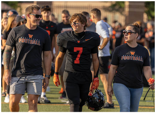 A person wearing a black football uniform with the number seven walks beside another person in a black Westmont football t-shirt and sunglasses. Description generated by AI
