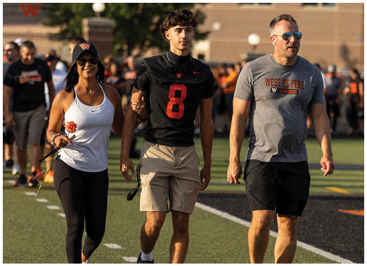 Two adults walk side by side on a field, one wearing a black sports jersey with the number 8 and beige pants, and the other wearing a gray West De Pere shirt and black pants. Description generated by AI
