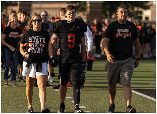Two people are walking side by side on a sports field, one wearing a black football uniform with the number nine and holding a helmet, and the other wearing a black t-shirt and gray shorts. Description generated by AI