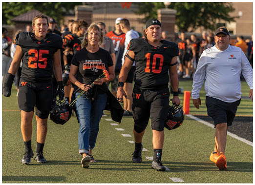 A person in an orange shirt and black shorts is reaching out to catch a football on a sports field while another person in black is diving toward them and a group of people stand in the background watching. Description generated by AI