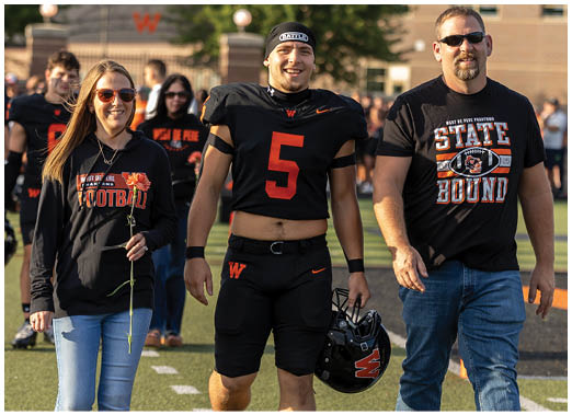 Two adults stand side by side outdoors, one wearing a black sports uniform with the number five and the other wearing a black t-shirt with a “State Bound" graphic. Description generated by AI
