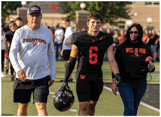 A person wearing a black football uniform with the number six and black face paint stands next to another person in a black sweatshirt with orange text that reads West De Pere. Description generated by AI