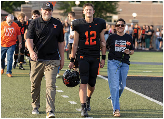 An adult in a football uniform holding a helmet walks arm-in-arm with two adults on a sports field, one holding a flower and wearing sunglasses. Description generated by AI