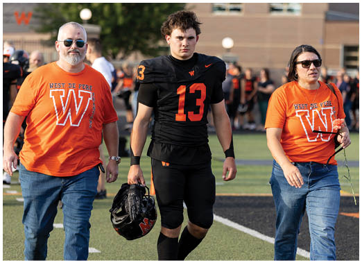 Three adults walk together on a sports field with one wearing a black football uniform holding a helmet and the other two wearing matching orange West De Pere T-shirts. Description generated by AI
