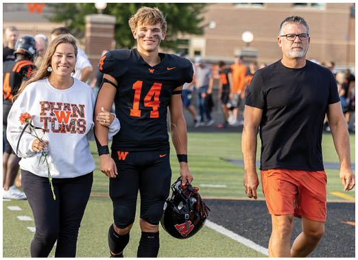 An adult wearing a sports jersey with the number 14 is walking arm-in-arm with another adult holding a flower on a sports field while a third adult walks beside them. Description generated by AI
