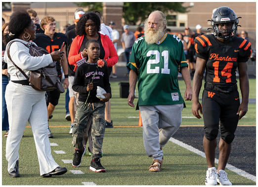 A group of people including a senior wearing a green sports jersey, a person in a black and orange football uniform, and others walking on a sports field during an event. Description generated by AI