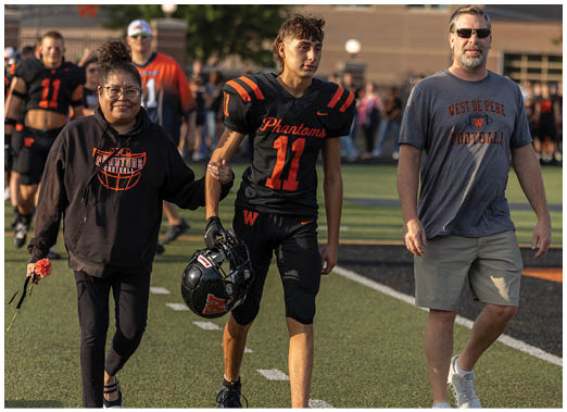 A person in a black football uniform with the number 11 and the word Phantoms walks beside another person wearing a gray West De Pere Football t-shirt and khaki shorts on a sports field. Description generated by AI