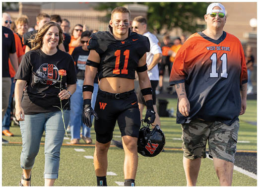 Two people wearing sports jerseys with the number 11 are standing outdoors, one in black athletic gear holding a helmet and the other in a black and orange jersey with camouflage pants. Description generated by AI