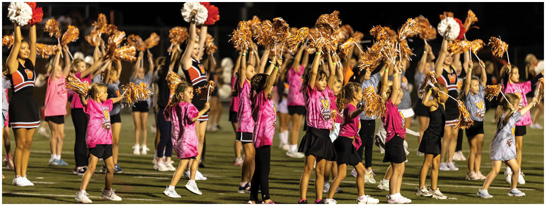 Children wearing matching pink shirts and holding orange pom-poms are running energetically on a sports field with adults in cheerleading outfits behind them. Description generated by AI