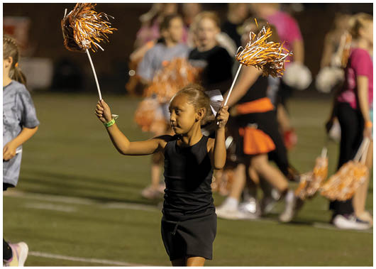 A child in a black sleeveless top with a white bow in their hair is holding an orange and white pom-pom while participating in a cheerleading activity on a field. Description generated by AI