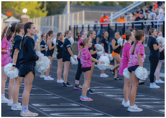 Children and adults dressed in athletic and cheerleading attire stand in rows on a track field holding white pom-poms during an outdoor event. Description generated by AI