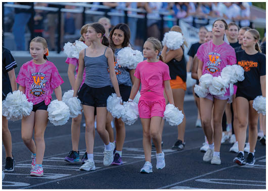Children wearing athletic clothing and holding white pom-poms are walking together on a paved surface. Description generated by AI