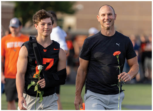 Two people are walking outdoors on a field, each holding a flower, with one wearing a sports jersey and an arm sling and the other wearing a casual athletic shirt. Description generated by AI