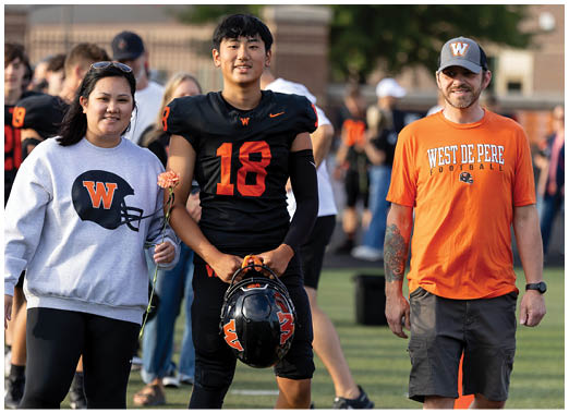 An adult wearing a football uniform stands between two adults, one holding a flower and the other wearing a West De Pere Football shirt and cap, on a sports field with people in the background. Description generated by AI