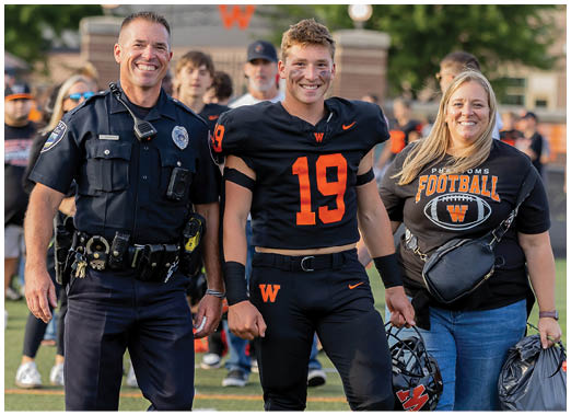 A person in a police uniform stands smiling next to a person in a black and orange football uniform and another person wearing a black football-themed shirt on a sports field with people in the background. Description generated by AI