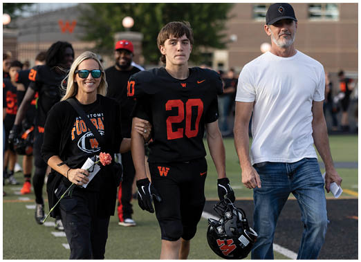 An adult on each side walks with a young person in a football uniform holding a helmet on a sports field during a daytime event. Description generated by AI