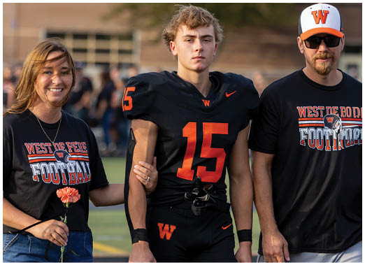 Three people stand together on a sports field with two wearing matching black shirts that say “West De Pere Football" and the person in the middle wearing a black football uniform with the number 15. Description generated by AI