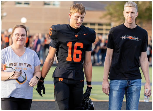 A person wearing a black football uniform with the number 16 is walking on a field holding hands with two people dressed casually, one in a gray West De Pere shirt and the other in a black shirt and jeans. Description generated by AI
