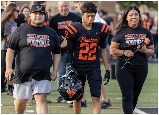 A person wearing a black and orange football uniform with the number 22 and the word Phantoms is holding a helmet while standing next to another person in a black West Deer Football shirt holding a flower. Description generated by AI